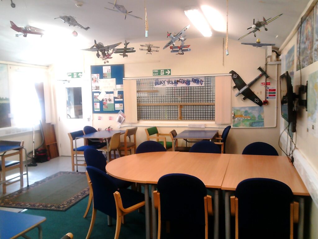Photograph of the 'Lobster Pot' craft and hobbies room. There are tables and chairs of various sizes arranged around the room, decorative posters and information displays on the walls, and a number of model aircraft suspended from the ceiling.