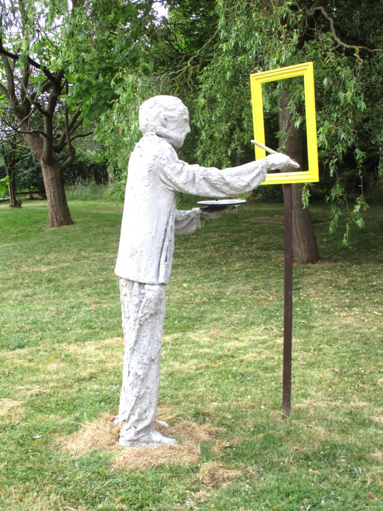 Photograph of a decorative statue in the grounds in the form of a man painting at an easel. It is cast in white stone.