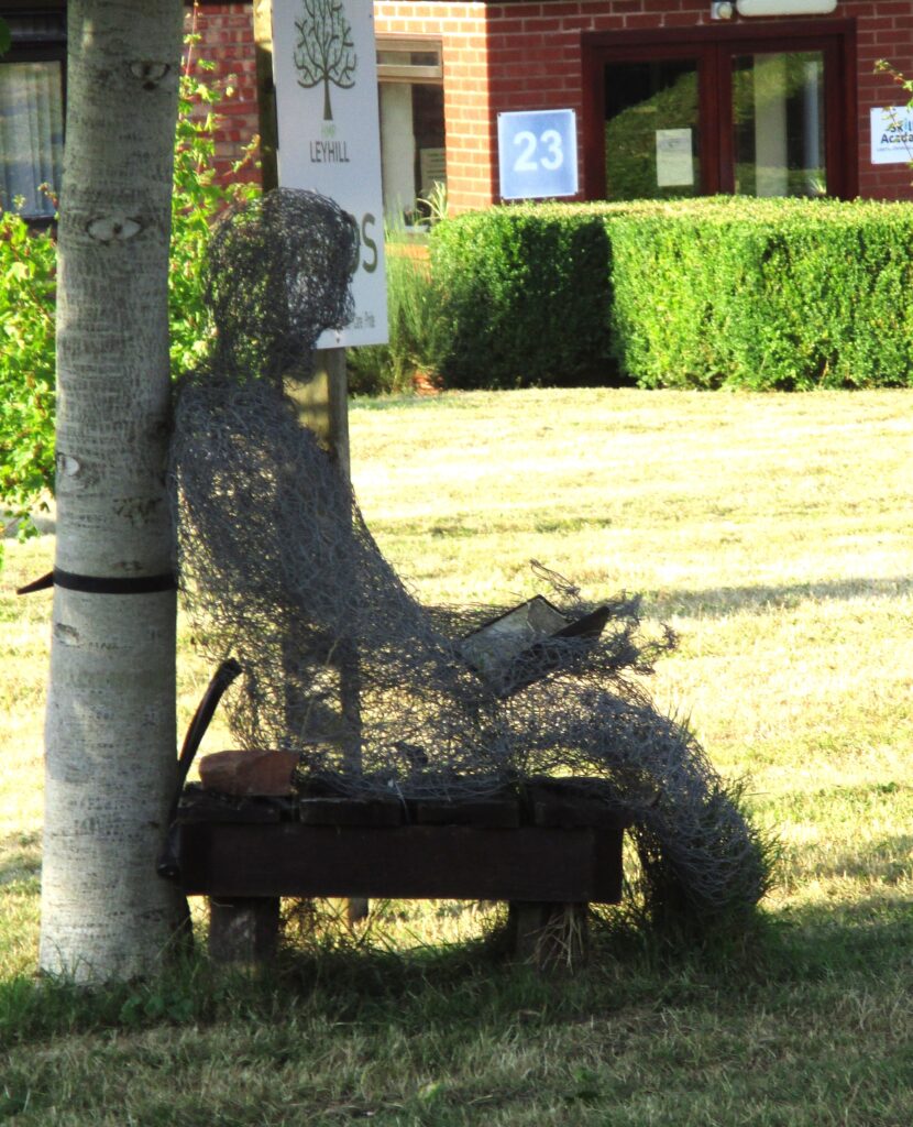 Photograph of a wire sculpture in the grounds. It depicts a man seating on a bench leaning against a tree.