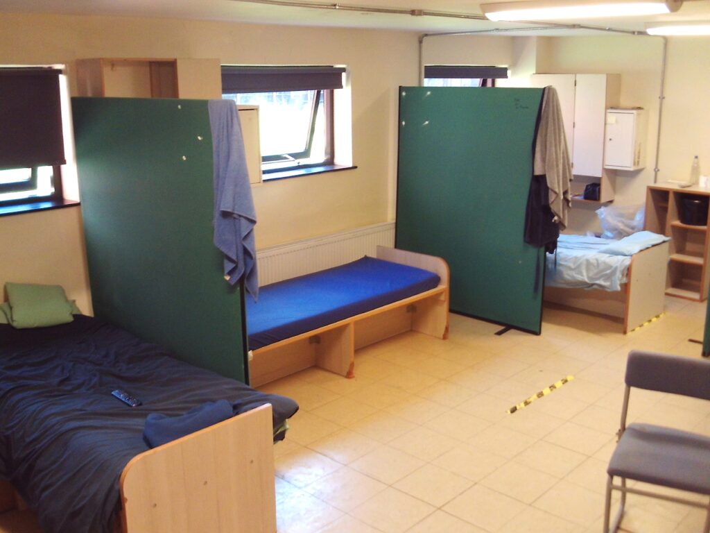 Photograph of the shared dormitory for new arrivals, showing single beds arranged around the edge of a hut, with partitions between them.