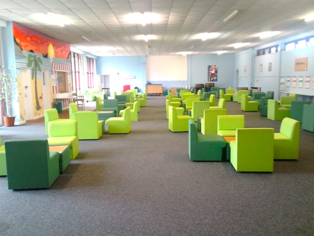 Photograph of the visits hall, with green chairs clustered around low tables arranged in rows down the length of the room.