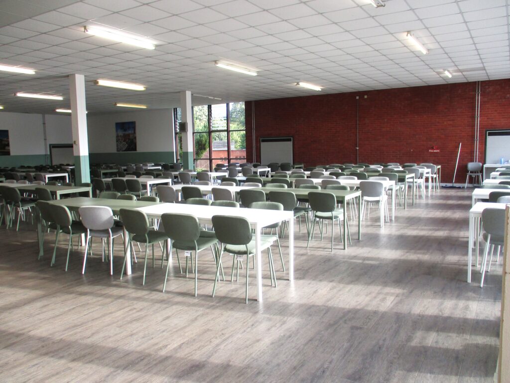 Photograph of the dining hall, a large, light room with rows of green chairs and white tables stretching across it.