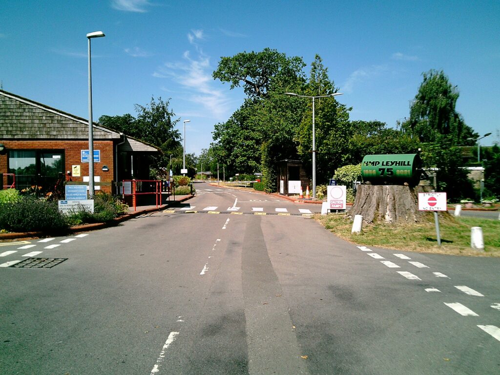 Photograph of the gatehouse and the road leading up to it at HMP Leyhill. The gatehouse building is on the left side of the road, while on the right are trees and the sign with the name of the prison on it.