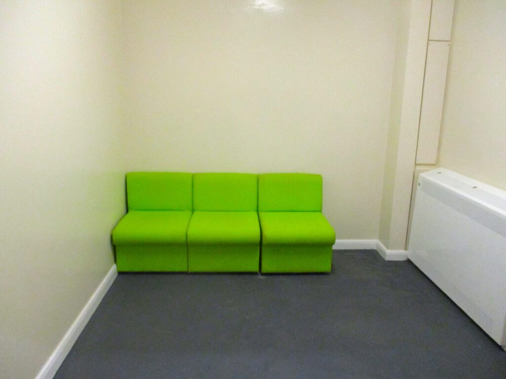 Photograph of the holding room, a small space with some seats covered in lime green fabric against one wall, the walls painted an off-white colour, and a grey carpet.