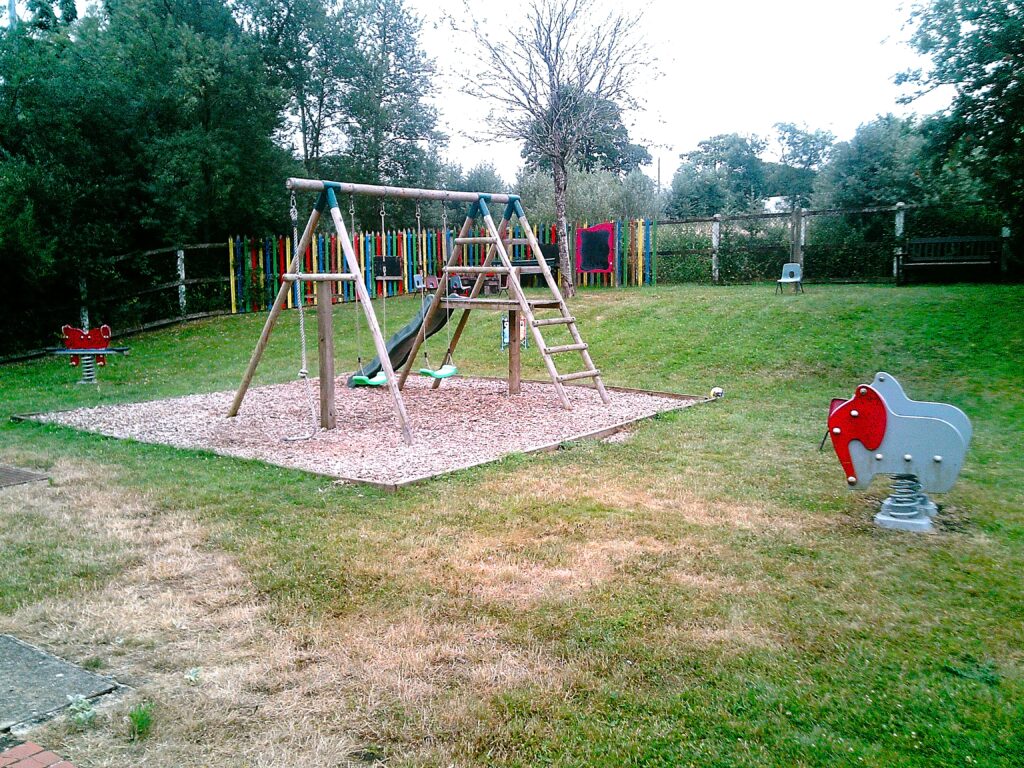 Photograph of the outside play area, a grass area on which several pieces of play equipment have been installed. they include a climbing frame with a swing and slide.
