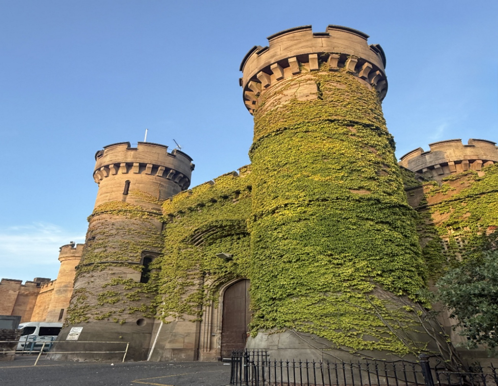 Photograph of HMP Leicester's external entrance, with a stone fortress frontage that looks like a castle. Round towers with crenels are partially covered in green ivy and set against a clear blue sky. There is a central wooden door, and a surrounding black metal fence, highlighting its medieval architectural style.