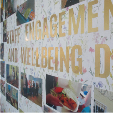 Photograph of a decorative wall display for Staff Engagement and Wellbeing Day featuring large gold letters on floral wallpaper. Surrounding the text are various small photos depicting activities and events related to staff engagement and wellbeing.