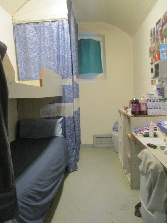 Photograph of a small, narrow well looked-after cell on 3s with a bunk bed on the left side and a cluttered desk on the right. The cell features blue curtains, a bunk bed with a muted blue bedspread, a window and various personal items on a desk.