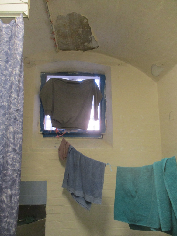 Photograph of a small, high window in a cell with peeling paint on ceiling and walls. A prison sweatshirt is being used as makeshift curtain and clothes, including a shirt and towels, hang on a line and over a rod beneath window.