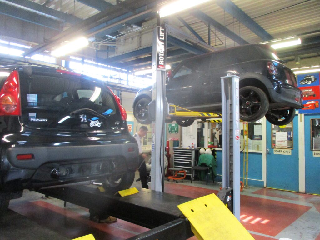Photograph of the Toyota workshop. Two cars can be seen hoisted up on hydraulic lifts to be worked on. Racks of tools can be seen in the background.