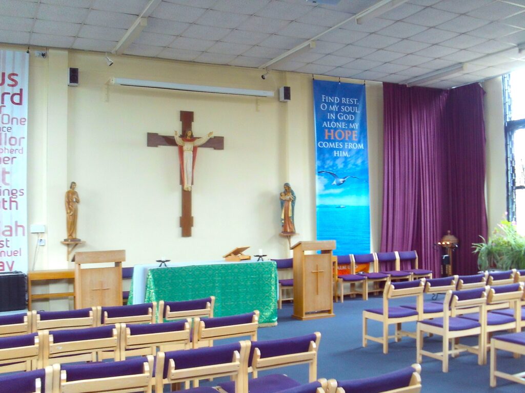 Photograph of the chaplaincy. A large cross is mounted on the wall behind the altar table, which has lecterns to either side. There are graphic panels with slogans on the wall. Ropws of chairs, with a central aisle, face them.