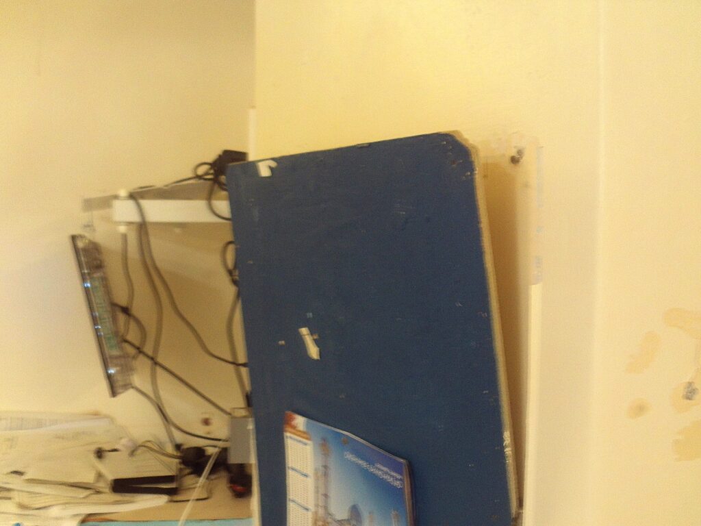 Photograph of a damaged fitting in a prison cell. A shelf and noticeboard are peeling away from a wall.
