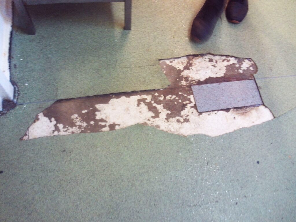 Photograph of damaged flooring on the servery floor on K wing. It shows a sizeable area where the floor covering has been torn away, exposing the surface underneath.