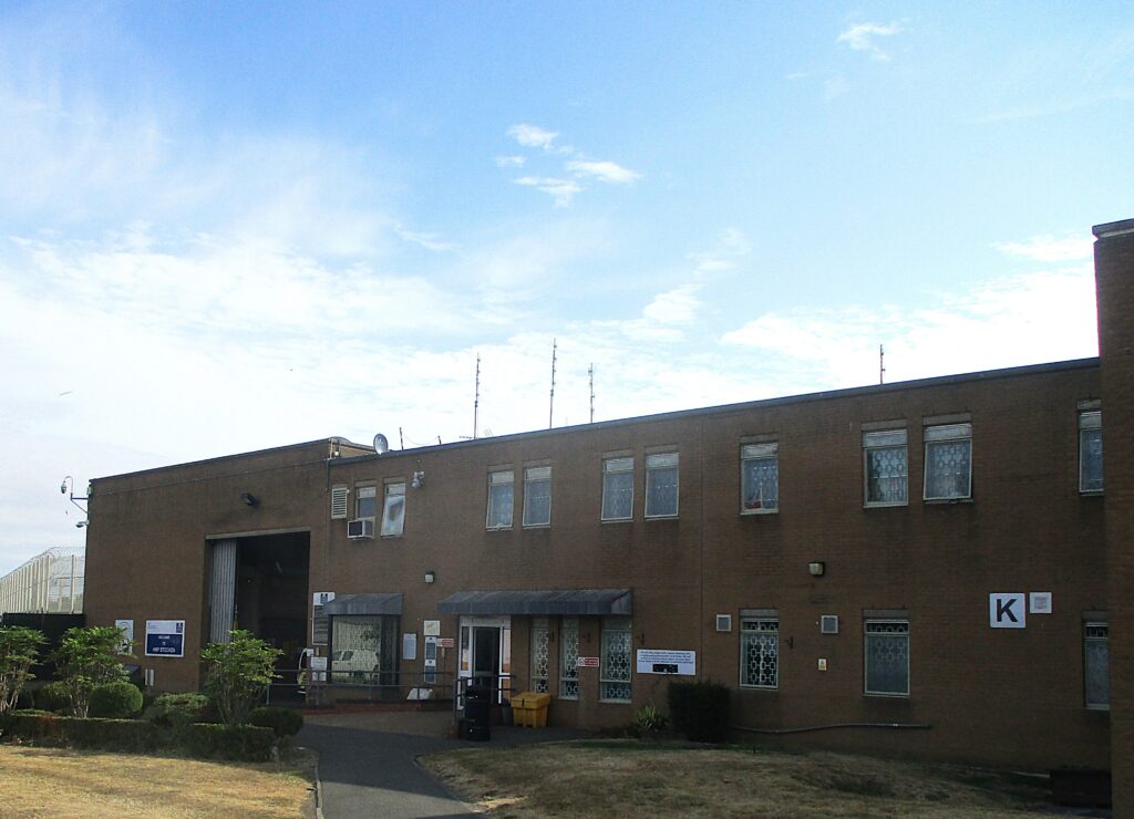 Photograph of the exterior of one of the buildings at HMP Stocken. It is a two-storey construction of brick, with windows visible on both floors. There is a large loading bay door at one end of the building and a small area of grass and shrubs is visible in front of it.