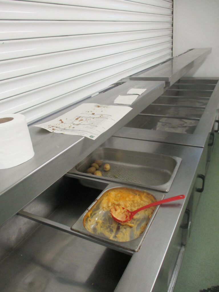 Photograph of a food servery area on M wing in which two trays containing food have been left overnight. One has a serving spoon still in it.
