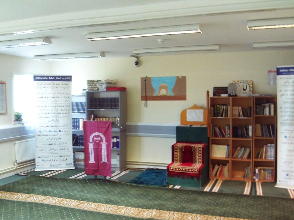 Photograph of the multi-faith room. The carpeted room has a bookcase and religious imagery arranged along the back wall.