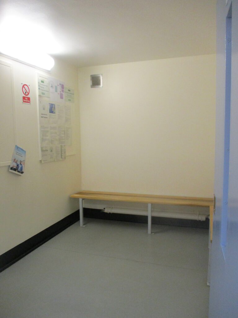 Photograph of the reception holding room. There is a wooden slatted bench against one wall and some displays of information on another.