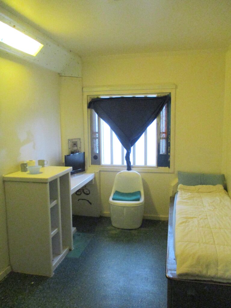 Photograph of a segregation unit cell. There is a bed against one side wall, a moulded plastic chair under the window, and a desk and storage furniture on the other side of the room. There is a small television on the desk.