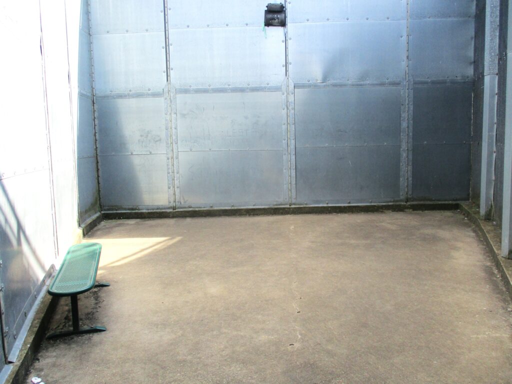 Photograph of the segregation unit yard, a small concrete space with a high fence around it. on the left side there is a small bench.