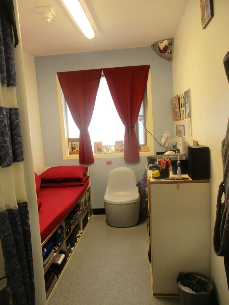 photograph of a single cell on O wing. There is a bed on one side, with storage underneath it, a plastic chair underneath it, and storage furniture on the other side of the room. The occupants things can be seen in the room.