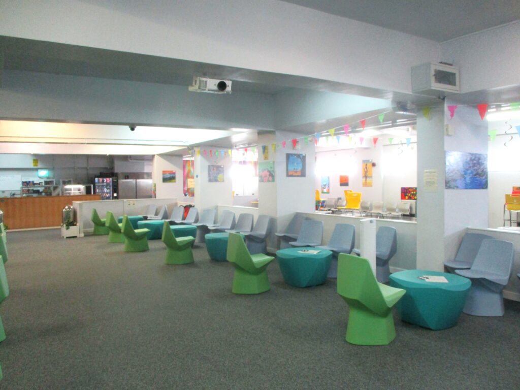 Photograph of the visits hall. Plastic moulded chairs and tables in shades of blue and green can be seen down one side of the room. there are displays and posters on the walls and a shop counter at the far end.