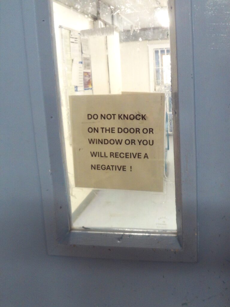 Photograph of a sign stick on the glass panel of a door warning prisoners that knocking on the door or window will result in them receiving a negative.