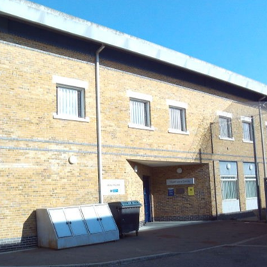 Photograph of the brick health care building's external entrance, showing white-framed windows, a covered doorway, roof overhang, wall signs and bins.