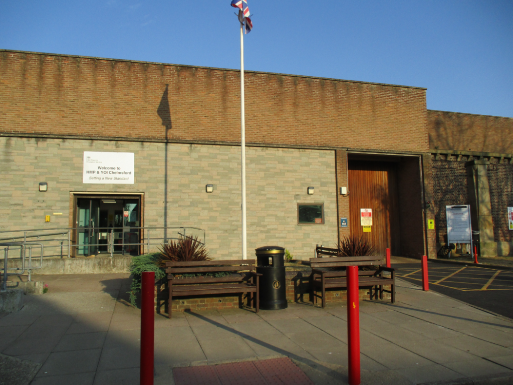 Photograph showing exterior entrance of HMP & YOI Chelmsford. There are signs, a flagpole, benches, a black waste bin, red safety bollards, an accessible ramp, and a blue-sky.