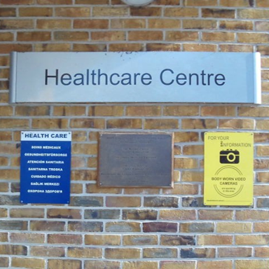 Photograph of a brick-wall entrance marked 'Healthcare Centre.' Blue healthcare notice, yellow CCTV-camera warning, and dark rectangular panel appear beneath sign.