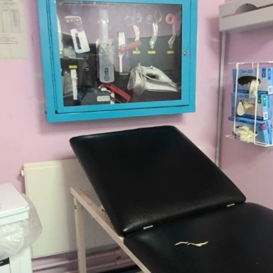 Photograph of the health care clinical room with a black reclining examination chair against pink walls. There is a wall-mounted cabinet, disposable glove dispenser and covered waste bin.
