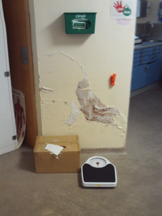 Photograph of the main health care clinical room, with damaged, peeling wall plaster, a cardboard box, and digital floor scale.