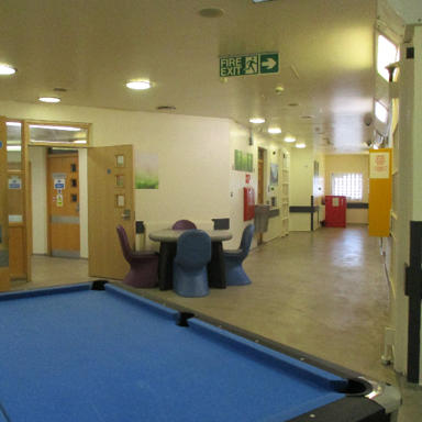 Photograph of an indoor recreational hallway in the ECU, showing a blue pool table, a table with seating, doors, and corridor extending toward a window.