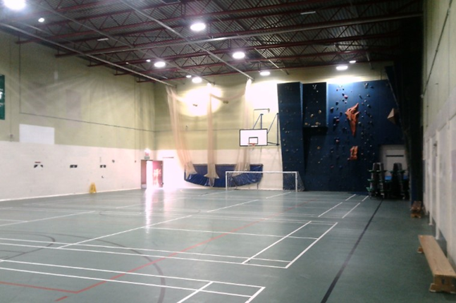 Photograph of an indoor multi-sport gymnasium featuring marked courts for basketball, football, and other activities. White and red court lines, basketball hoop, small soccer goals, blue climbing wall, benches, and overhead lighting define the layout.