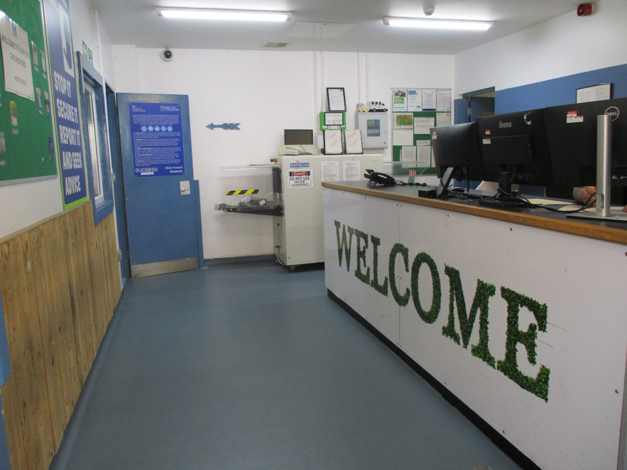 Photograph of a reception or information desk inside a clean facility, with multiple computer monitors, service counter, blue doors, and posted instructional signs. Large green &ldquo;WELCOME&rdquo; lettering decorates the desk front, while blue-and-green wall signage provides visitor guidance and facility information.