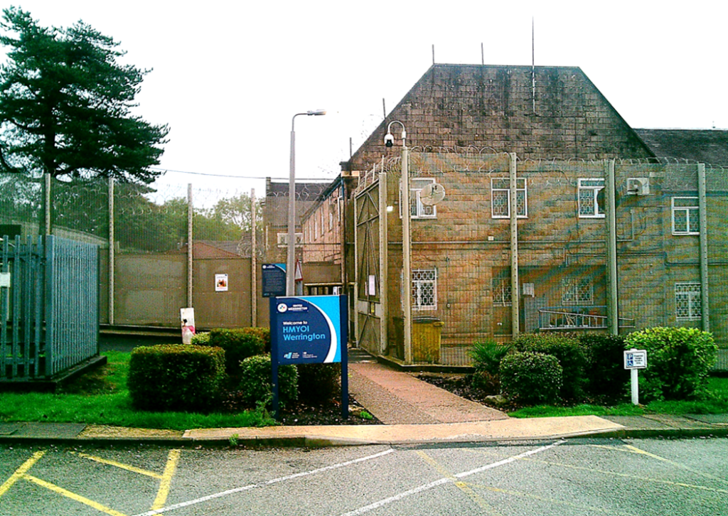 Photograph shows HMYOI Werrington, a brick youth offender institution building behind security fencing. Blue entrance sign, gated access, barred windows, CCTV, trimmed shrubs, street markings, and adjacent industrial structures.