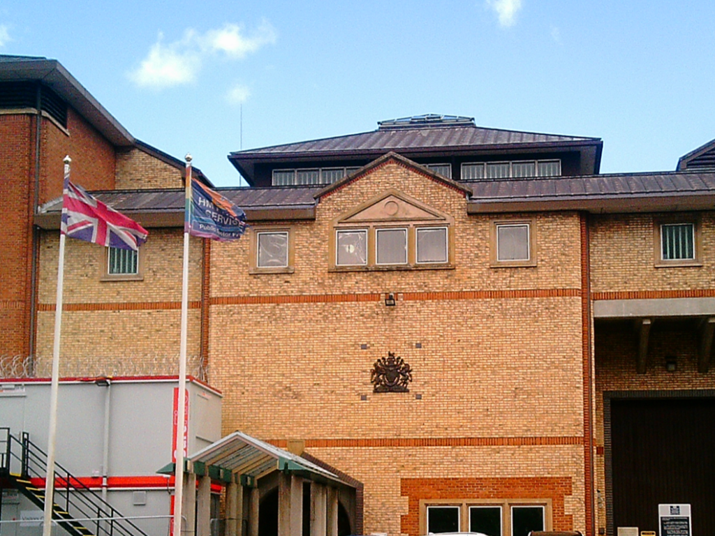 Photograph shows a large historic red-brick civic or institutional building beside an urban street. To the forefront, two flags on white poles, one a Union Jack and one a HMPPS service flag, an ornate central emblem and covered entrance.