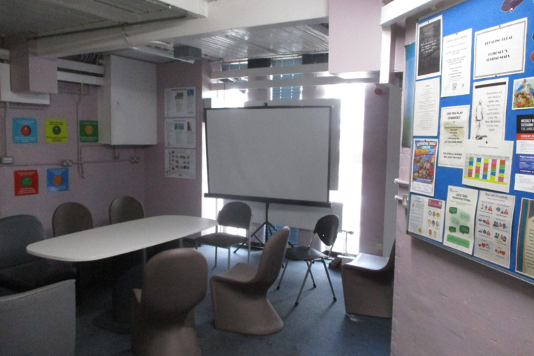 Photograph of a small induction room with oval table, chairs, projection screen, and bright windows. Right-side bulletin board displays colourful posters, charts, notices, and informational materials, while left wall features additional labelled panels.