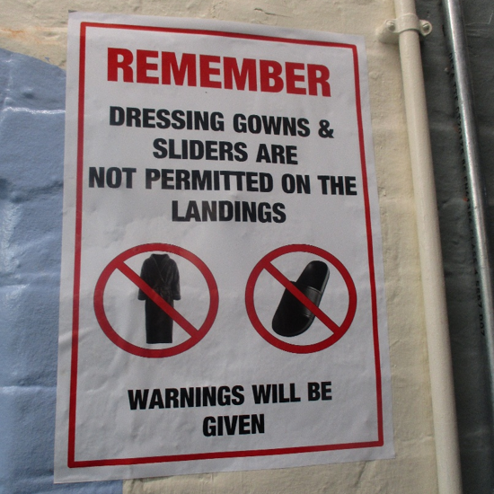 Photograph of a posted safety sign reminding visitors that dressing gowns and slippers are prohibited on landings. The red-bordered sign uses bold black and red text, crossed-out gown and slipper symbols, and warns that violations will receive warnings.