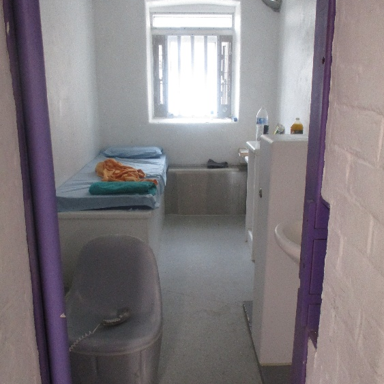 Photograph showing an occupied segregation cell viewed through a purple-painted doorway, showing a single bed with folded clothing on it, a white sink and storage.