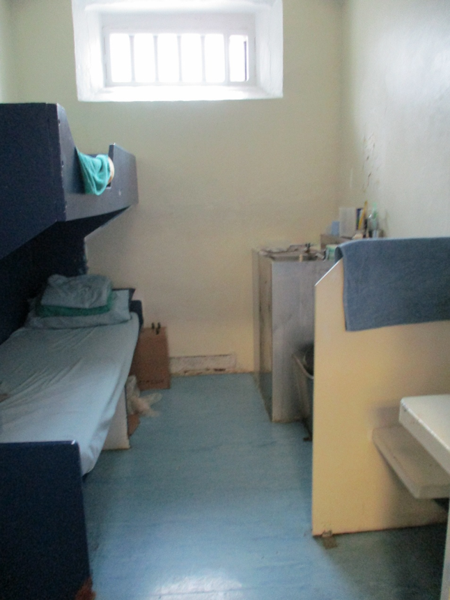 Photograph documents a narrow prison cell, with stacked bunk beds and a small barred window. Blue-grey bedding, pale walls, compact sink and toilet, cabinet, and sparse personal items emphasize confined living conditions.