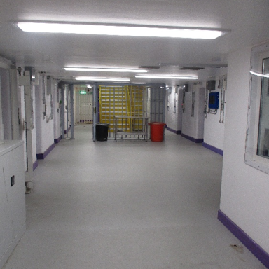 Photograph showing the brightly lit segregation unit corridor with white walls, grey flooring, purple baseboards, and multiple doors along both sides. Yellow stairs, black and red bins, fluorescent ceiling lights, and a green exit sign appear at corridor’s far end.