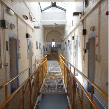 Photograph shows a prison wing landing/corridor with white brick walls and cell doors lining both sides. The landing overlooks another wing below, and has yellow metal safety railings and wire-mesh netting.