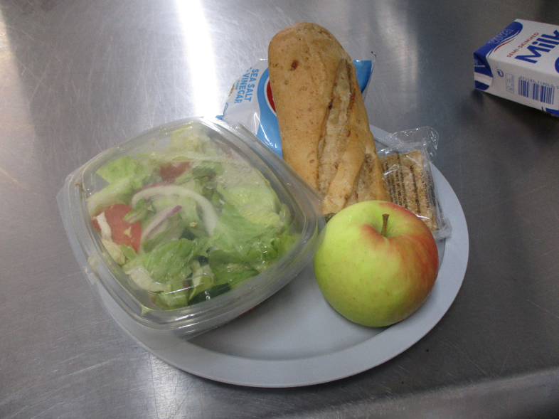 Photograph of a meal arranged on a plate, including a green salad in a clear plastic box, part of a baguette, a green-red apple, packaged snack, a packet of crisps and a milk carton.