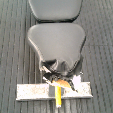 Photograph of a damaged black padded chair or seat. The material is torn and hardware exposed.