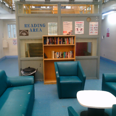 Photograph shows a reading area with teal sofas and armchairs, a white table, and a central wooden bookshelf. There is a blue “READING AREA” sign, books, informational posters and windows.