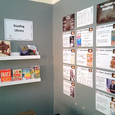 Photograph of a reading library corner with two white shelves displaying assorted books and magazines beneath a “Reading Library” sign. Gray wall features colourful “Power of Reading” posters, book recommendations and reading-related displays.