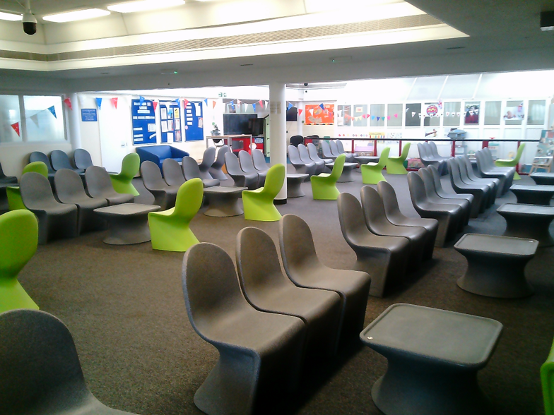 Photograph showing a visits hall lounge with rows of moulded chairs and small tables. Gray seating, bright lime-green chairs, blue information boards, bunting, and large windows create a colourful space.