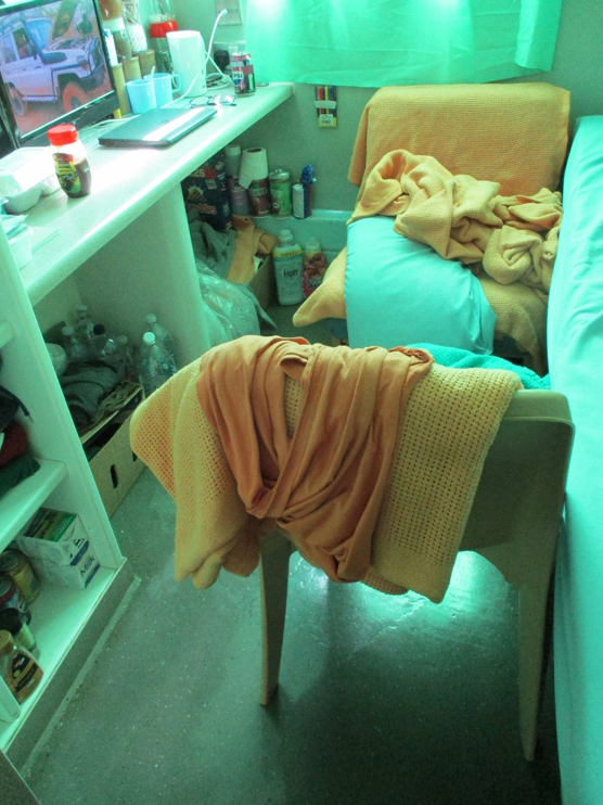 Photograph of a wheelchair user's cell, showing a cramped room with two chairs draped in orange blankets, a counter, shelves, and numerous bottles and personal-care supplies.