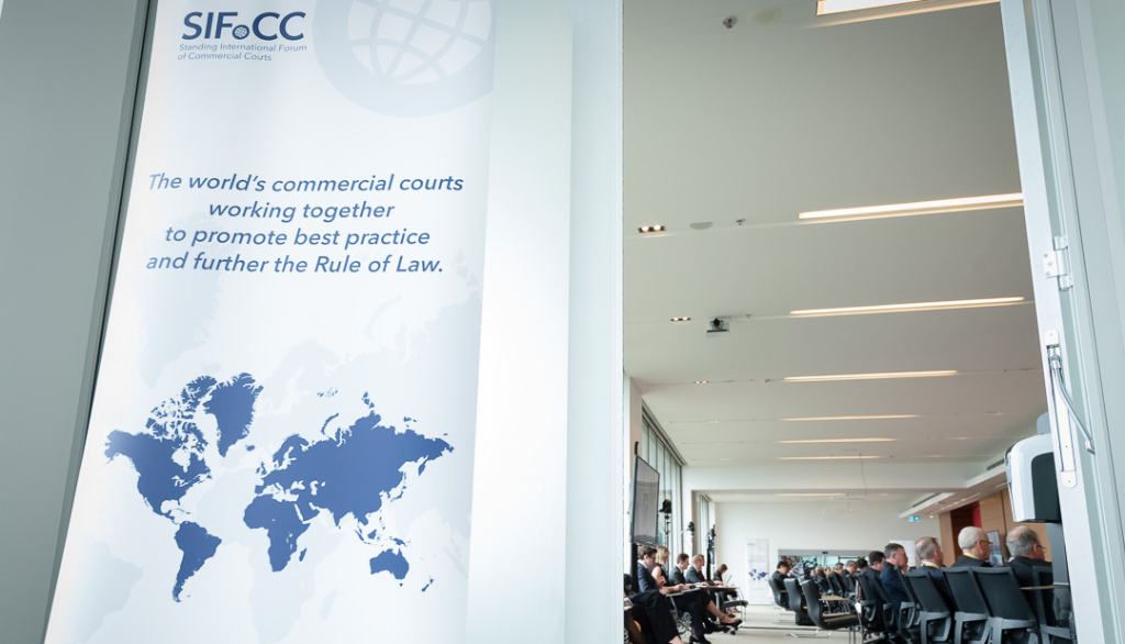 Image of a door open into a room of judges with a SIFoCC banner outside the door which reads 'The world's commercial courts working together to promote best practice and further the Rule of Law.
