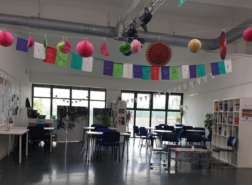 Photo showing an example of a community hub. The image features a room decorated with colourful paper decorations and banners hanging from the ceiling. The room has several tables and chairs, with shelves and large windows in the background.